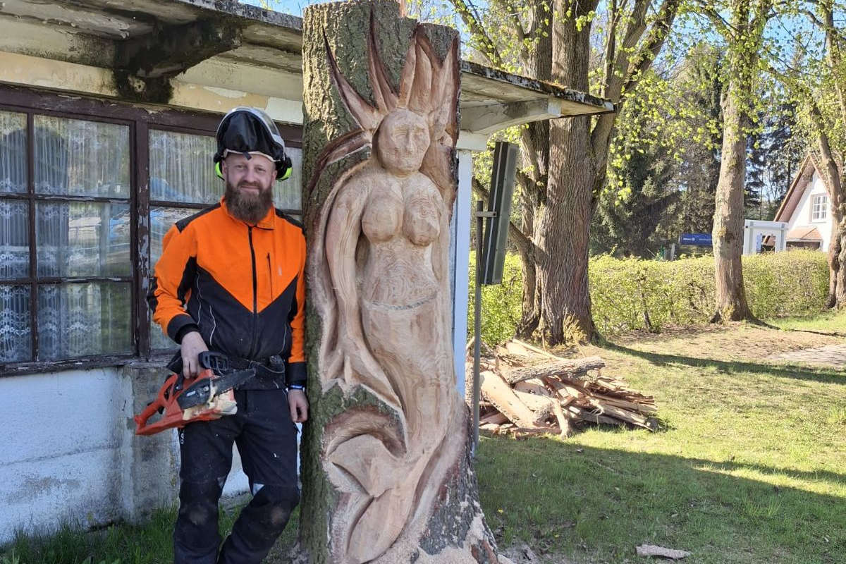 Ein Mann in Schutzkleidung steht neben einem geschnitzten Baumstamm, der eine detailreiche Skulptur einer Frau (Nixe) mit wallendem Haar zeigt. Er hält eine Kettensäge in der Hand und lächelt in die Kamera. Stolz repräsentiert er den Förderverein Bad Oppach in einer sonnigen Umgebung im Freien.