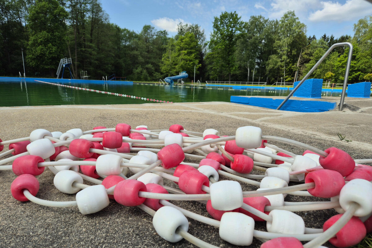 Ein Bündel weißer und rosafarbener Kunststoff-Bahnenbegrenzer liegt auf dem Beton neben dem Freibad Bad Oppach, umgeben von Bäumen unter einem teilweise bewölkten Himmel. Im Hintergrund sind blaue Beckenränder und eine Wasserrutsche zu sehen.