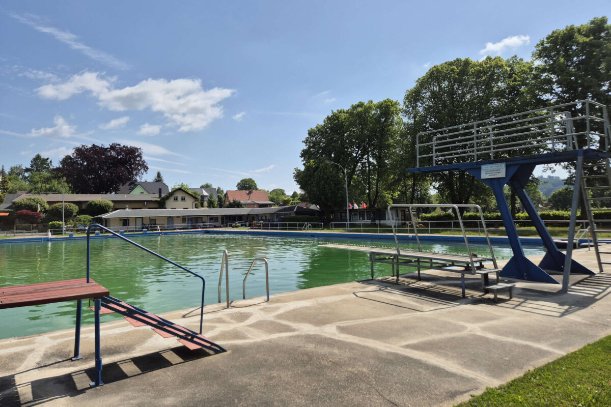 Das Freibad Oppach ist ein öffentliches Freibad mit grünlichem Wasser, umgeben von Bäumen und Häusern. Ein blauer Sprungturm und eine Metallleiter stechen hervor, während der Himmel über dem Freibad meist klar ist und nur wenige Wolken zu sehen sind.